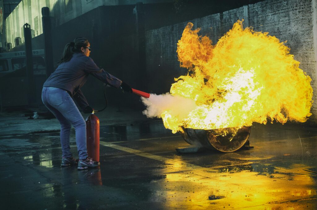 pexels-photo-571252-571252 A woman uses a fire extinguisher to put out a large flame in an outdoor setting, showcasing fire safety.